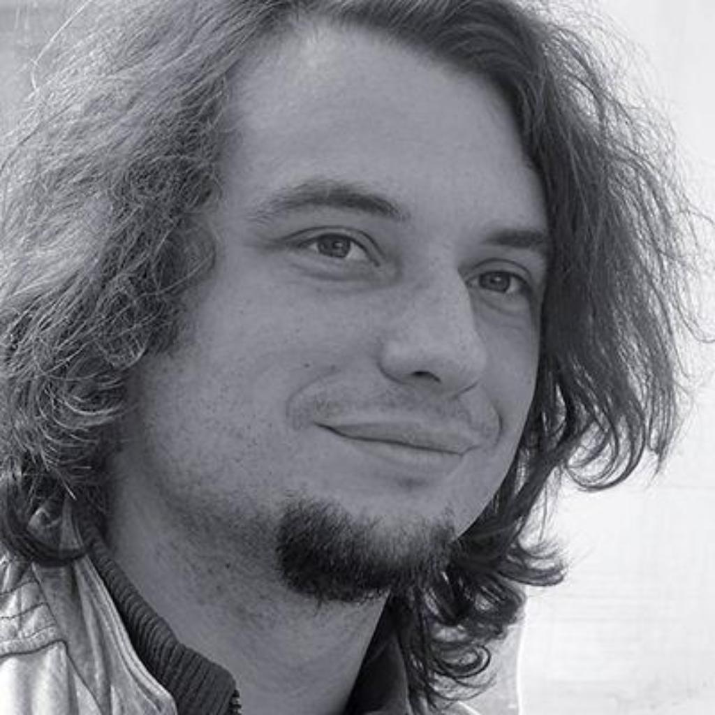 Porträt von Martin Fiedler mit schulterlangem Haar und Ziegenbart, leicht lächelnd und seitlich an der Kamera vorbeiblickend vor hellem, unscharfem Hintergrund